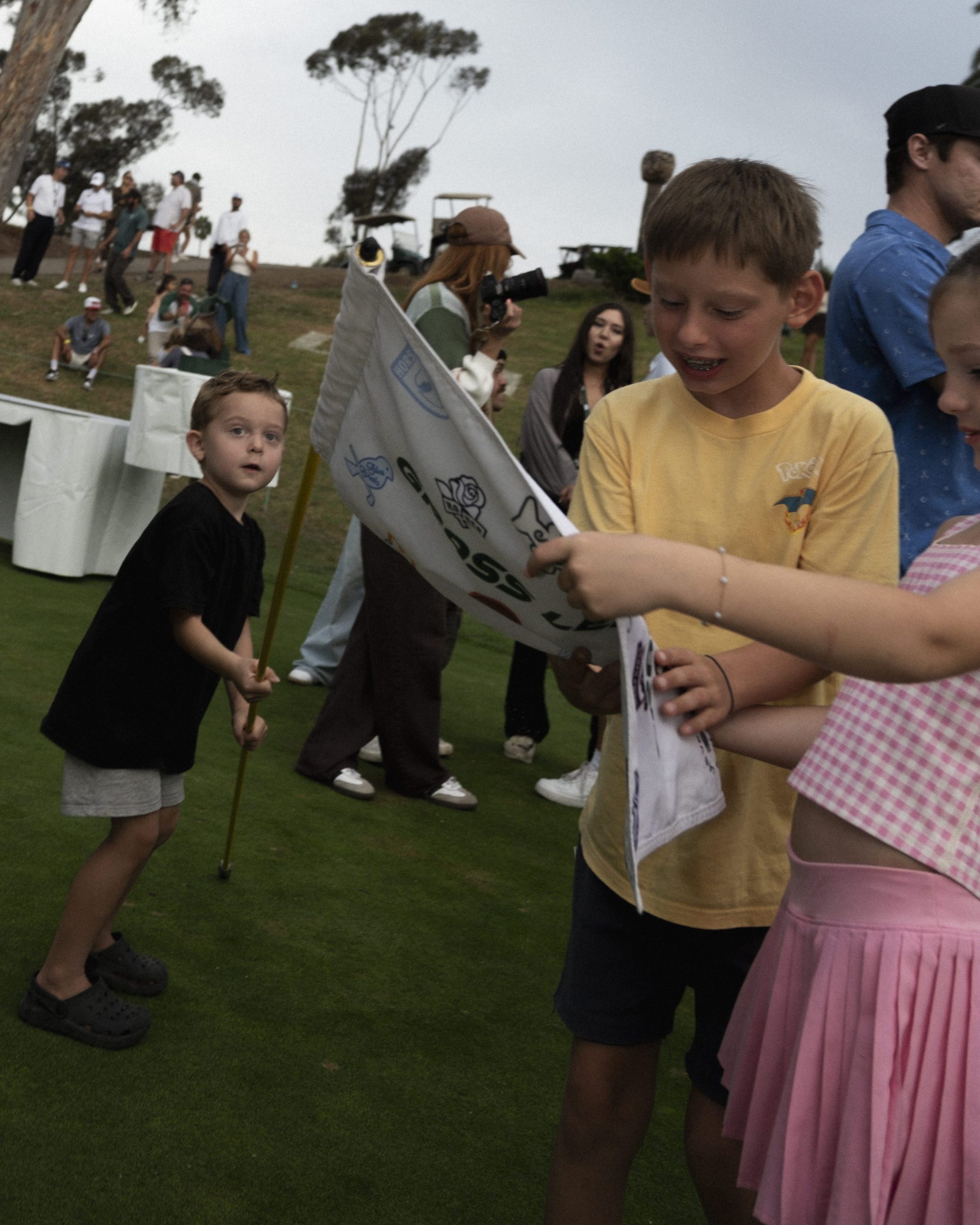 Kids holding a signed Grass League pin flag with fans on the hillside behind